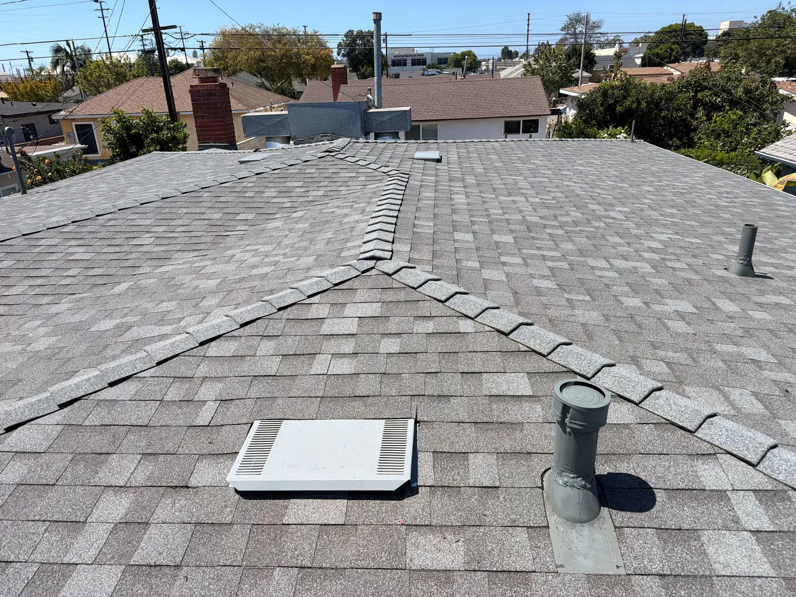 A gray asphalt shingle roof with multiple hip ridges, a white rectangular attic vent, and two pipe vents, overlooking a residential neighborhood.