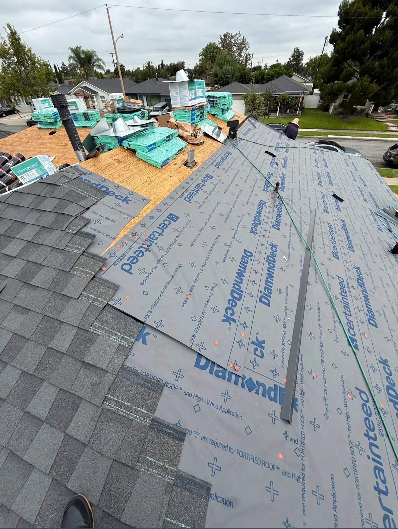 A residential roof under replacement with gray synthetic underlayment installed, partial shingle coverage on one section, and stacks of roofing materials and vents staged near the ridge.