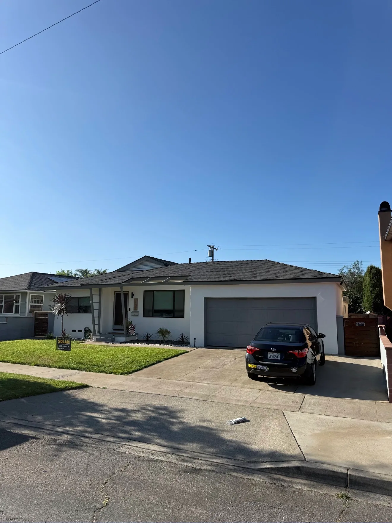 Single-story house with a dark shingle roof, white stucco exterior, and a black car parked in the driveway in front of a two-car garage.