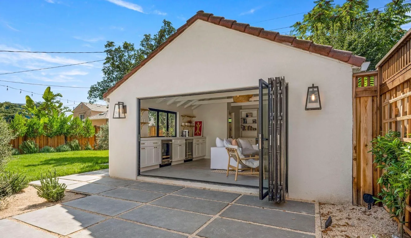 A detached structure with a terra-cotta tiled roof and open bi-fold glass doors revealing a furnished interior with a sofa, cabinetry, and shelving, set on a paver patio with a landscaped yard.