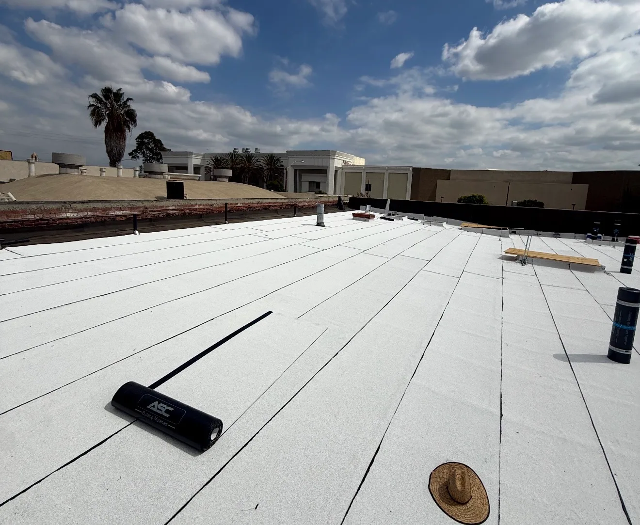 A flat commercial rooftop covered with light-colored cap sheet membrane, with a roofing roller tool, a roll of membrane material, a straw hat, roof vents, and surrounding buildings under a partly cloudy sky.