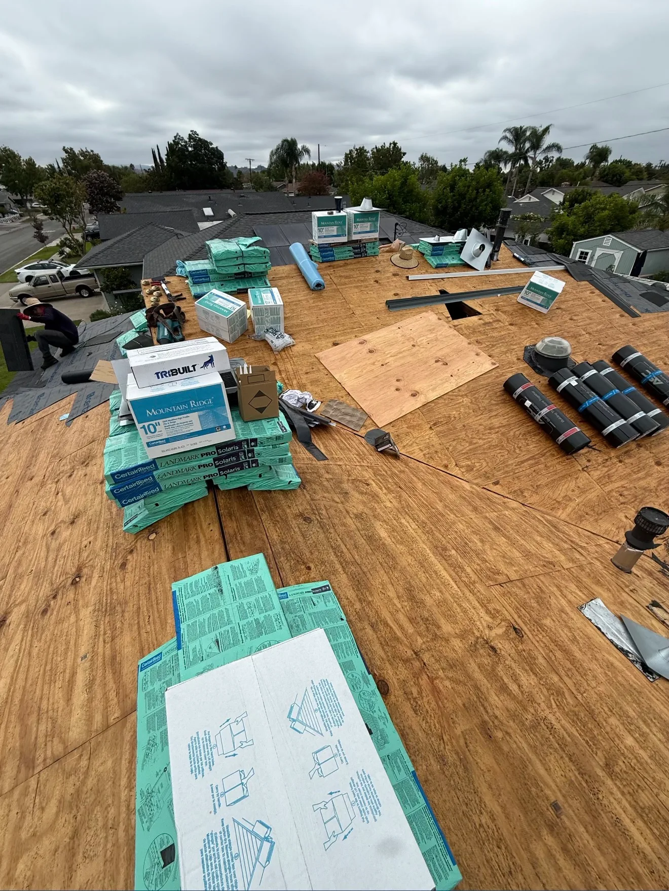 A roof under construction with exposed plywood decking, stacked bundles of shingles, boxes of ridge caps, rolls of underlayment, and a worker kneeling near the edge under an overcast sky.
